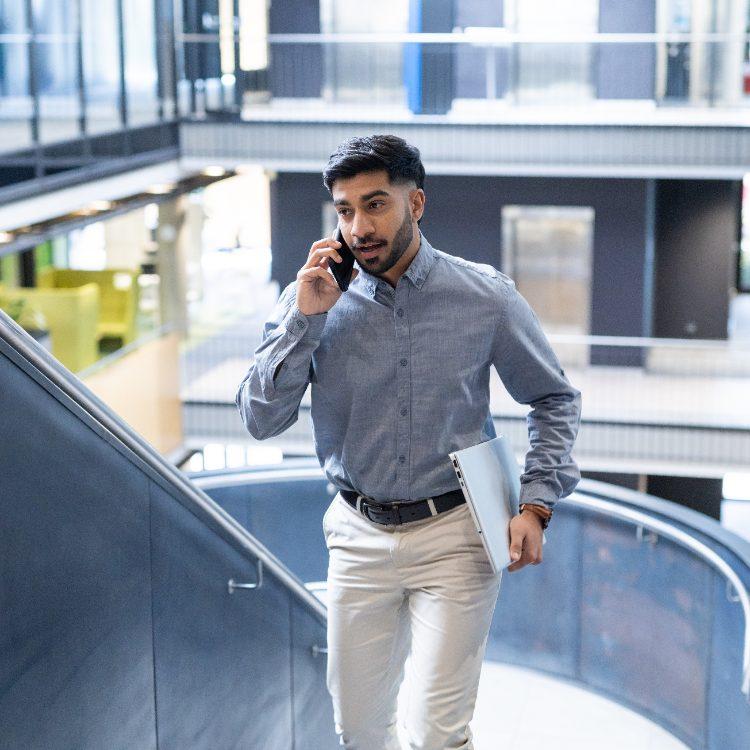 Man on a phone call walking up the stairs of an office atrium with a laptop