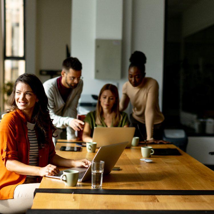 Smiling woman at a laptop while three colleagues review a screen at a long wooden table