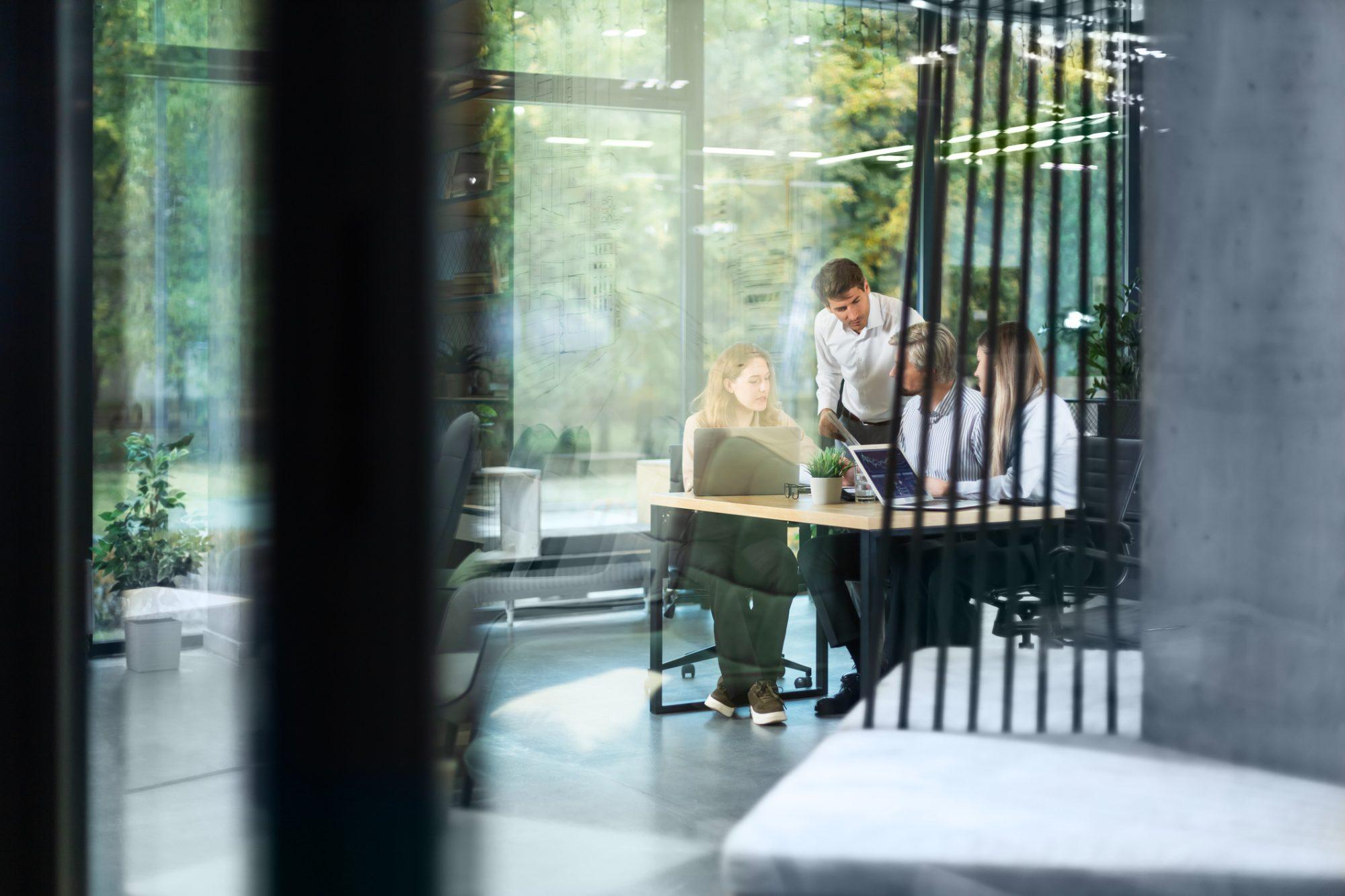 Four colleagues reviewing a laptop in a glass meeting room surrounded by greenery