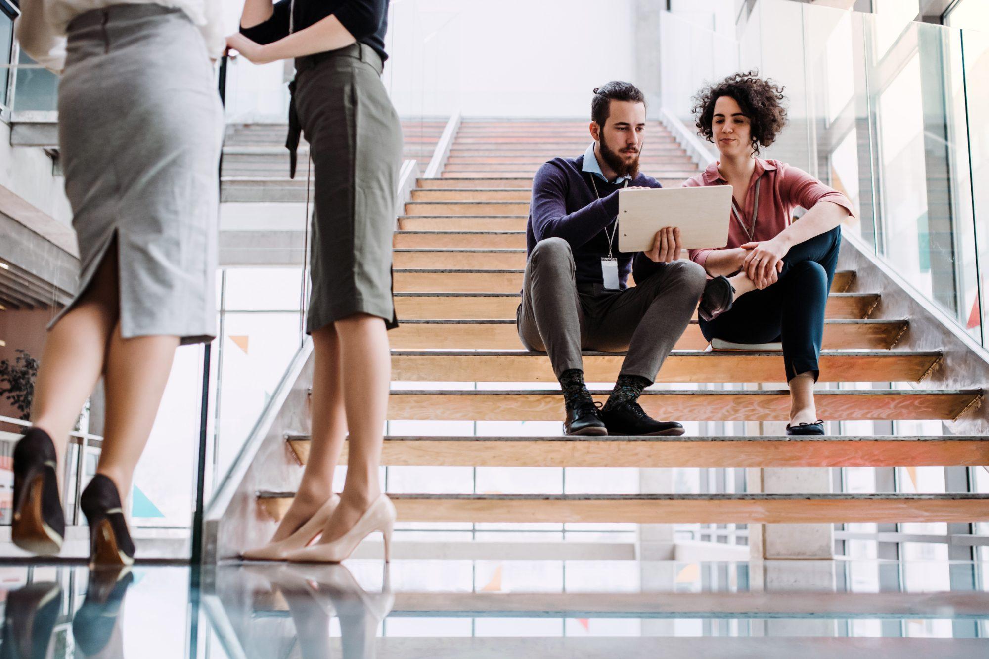 Two colleagues sitting on office stairs reviewing a clipboard