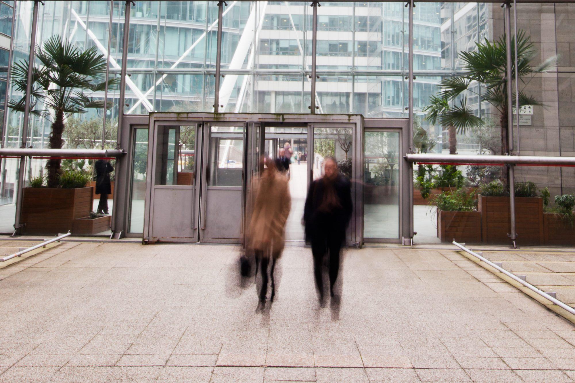 Two blurred figures walking out of a glass entrance beneath a steel-framed glass facade