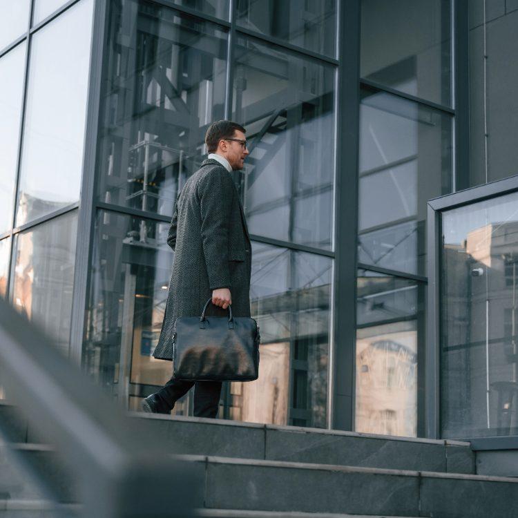 Man in a long coat carrying a briefcase up the steps of a glass office building