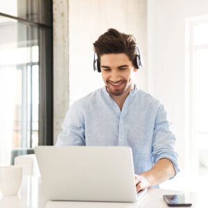 Male employee wearing headphones smiles down at his laptop computer while working in the office.