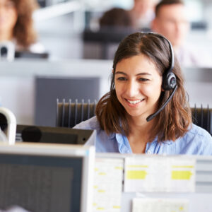 Female employee smiling while wearing a phone headset and looking at her computer monitor while using Microsoft Teams Contact Center.