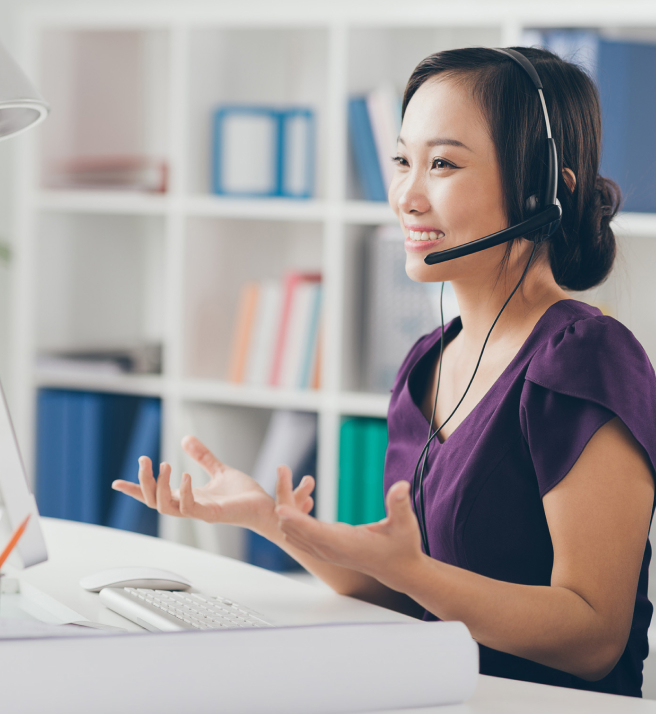 Woman in a purple blouse, wearing a headset, smiling and gesturing while on a video call. She sits at a desk with a computer and a bookshelf behind her.