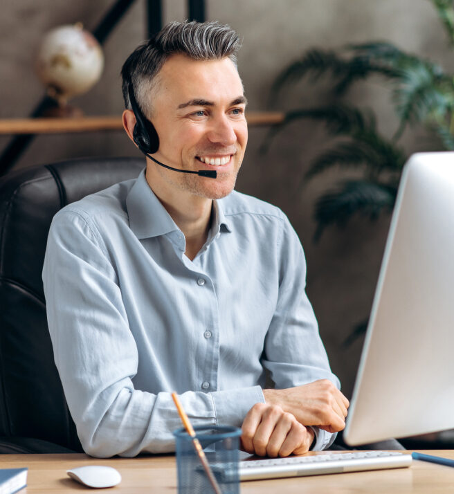 Smiling man with a headset sitting at a desk, engaging with his computer. The background features a globe and green foliage, suggesting an office setting.