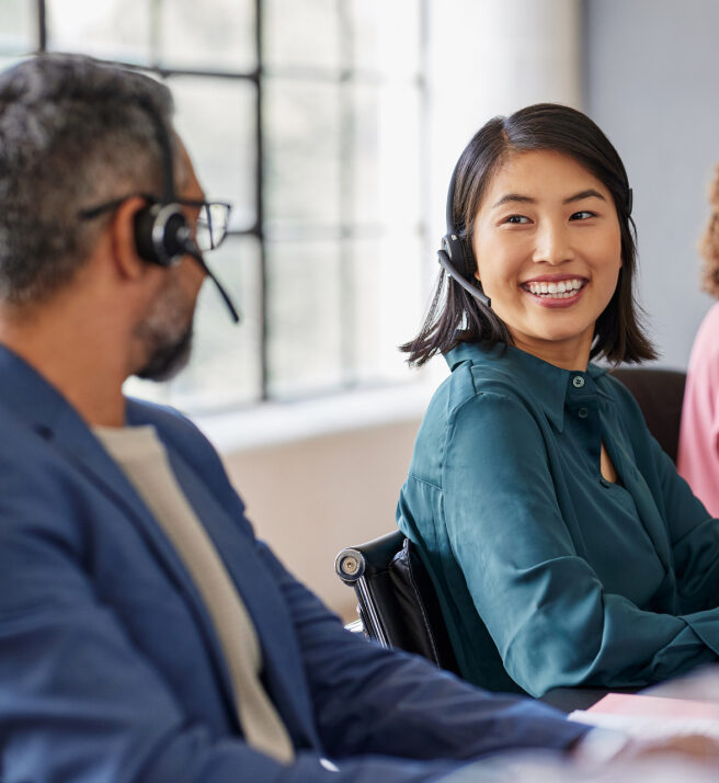 A woman in a teal blouse and headset smiles at a man in glasses wearing a headset. They appear to be in an office, suggesting a friendly workplace.
