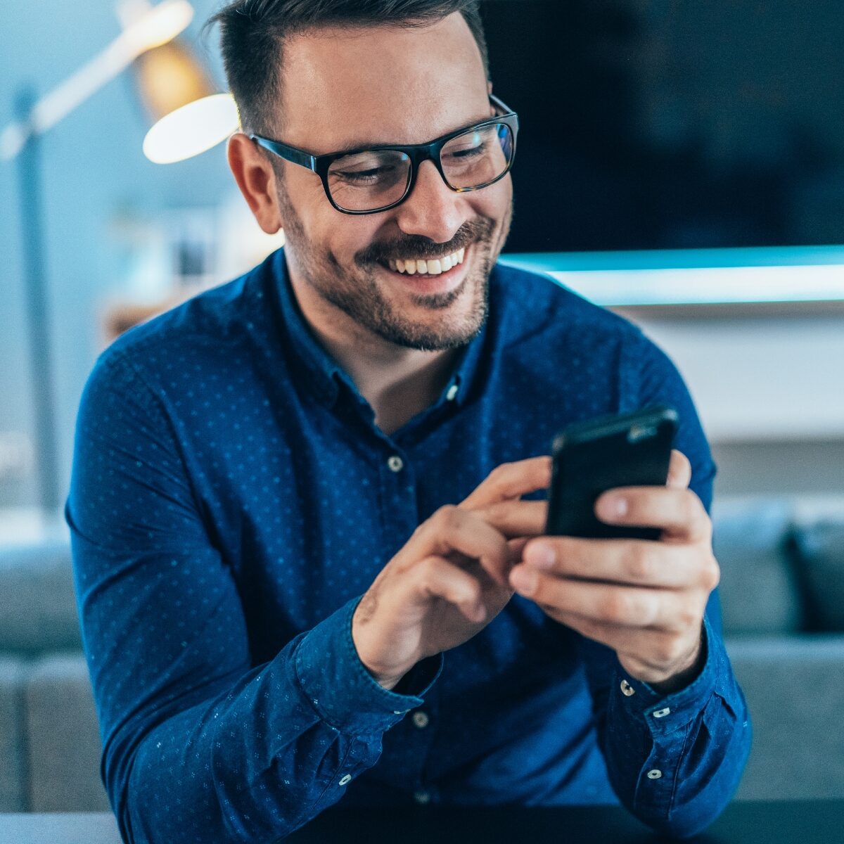 A man wearing glasses and a blue shirt smiles as he looks at his phone, sitting in a cozy room with soft lighting. The mood is cheerful and relaxed.
