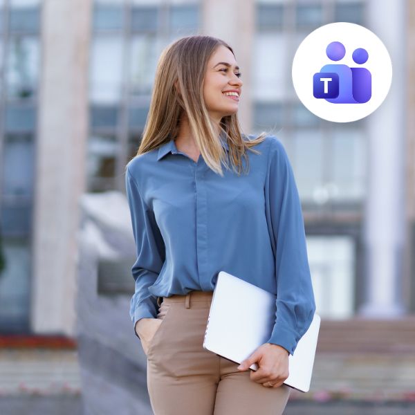 A woman in a blue shirt and beige pants, smiling while holding a laptop outside a modern building. Microsoft Teams logo is in the top right corner.