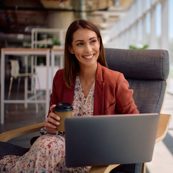 Smiling woman in a patterned dress and red blazer holds a coffee cup while using a laptop in a modern, well-lit office, conveying a positive and focused mood.