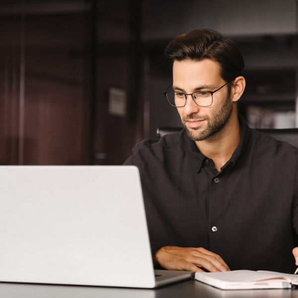 A man with glasses and short dark hair types on a laptop in a dimly lit office. He wears a black shirt, conveying focus and concentration.