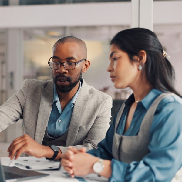 A focused professional duo in an office discusses work over a laptop. The man gestures while explaining, and the woman listens attentively, conveying teamwork.