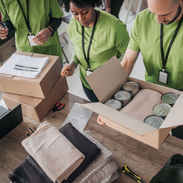 Volunteers in green shirts pack canned goods and blankets into boxes on a wooden table, creating a sense of teamwork and community support.