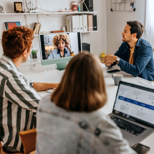 Three people at a table engage in a video call with a person on a computer screen. The setting feels collaborative and professional.
