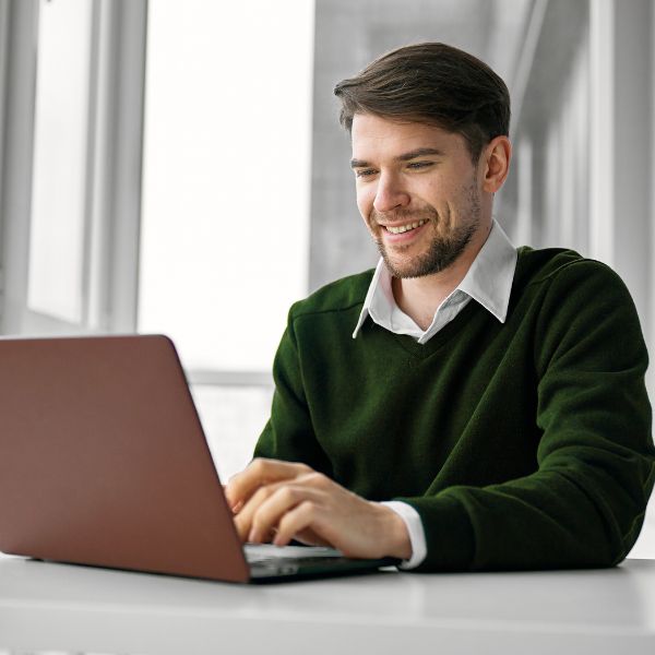 A smiling man in a green sweater types on a laptop at a bright, sunlit desk. He appears focused and content, suggesting a pleasant work moment.