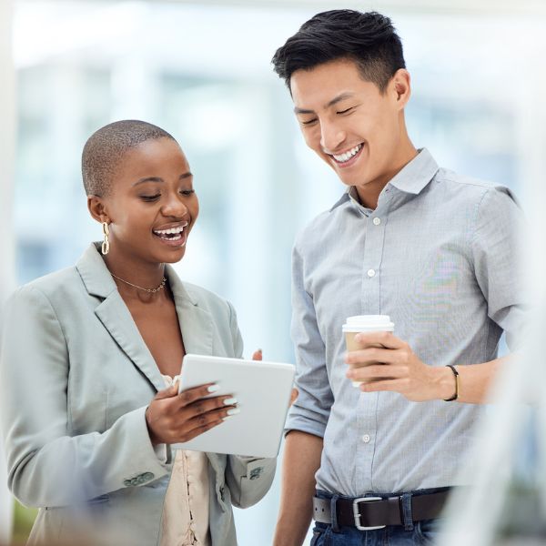 Two people standing indoors, smiling and looking at a tablet. One is holding the tablet, and the other is holding a takeaway coffee cup.