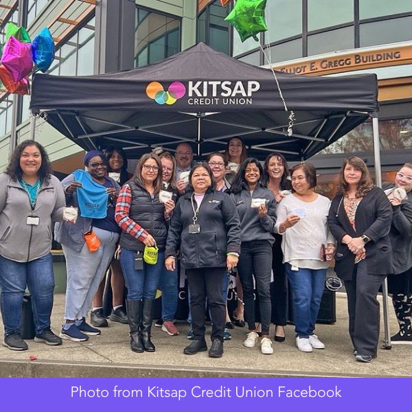 A group of people pose and smile in front of a Kitsap Credit Union tent outside a building, holding small items, with colorful balloons in the background.