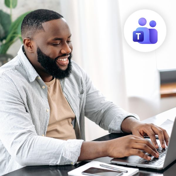 A smiling man works on a laptop in a bright room, wearing a striped shirt. The Microsoft Teams logo floats nearby, indicating collaboration.