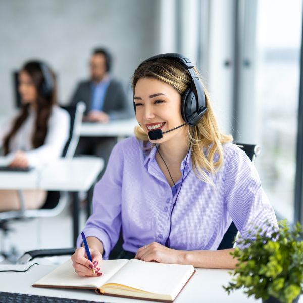 Smiling woman in a call center wearing a headset, taking notes in a notebook. Bright office setting with colleagues blurred in the background.