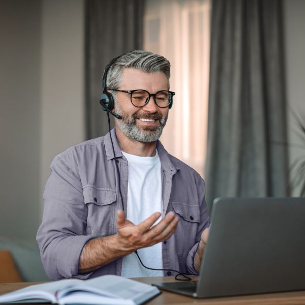 Smiling man with glasses and headphones, participating in a video call on a laptop. He gestures expressively, conveying a welcoming and engaged tone.