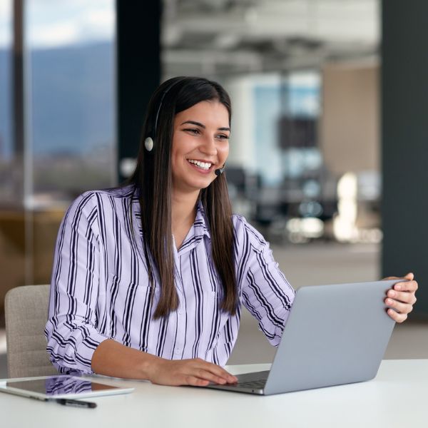 Smiling woman in a striped shirt wearing a headset, using a laptop in a bright office. She exudes a friendly and professional demeanor.