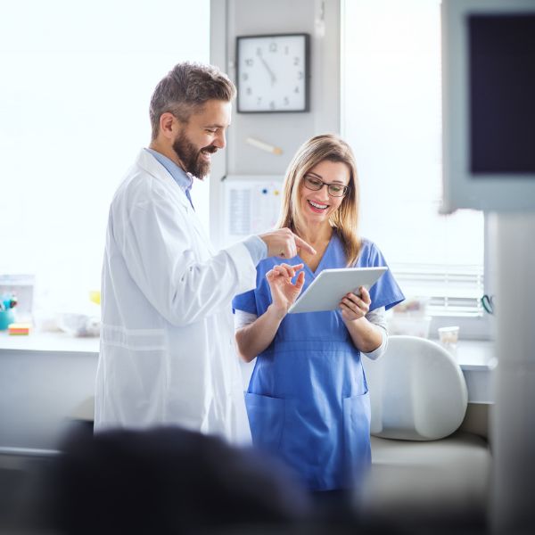 A doctor and a nurse smile while reviewing a tablet in a bright hospital room. The clock and medical equipment create a professional, collaborative tone.