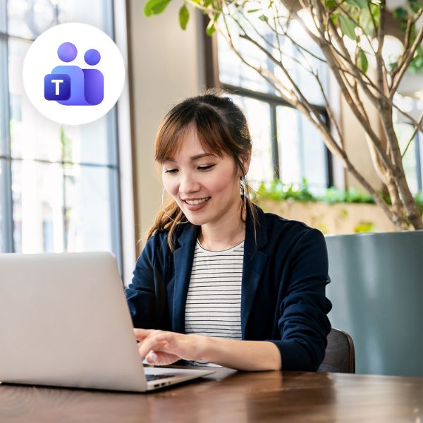 Woman smiling while using a laptop in a bright office, with a Microsoft Teams logo in the corner. She appears focused and engaged.