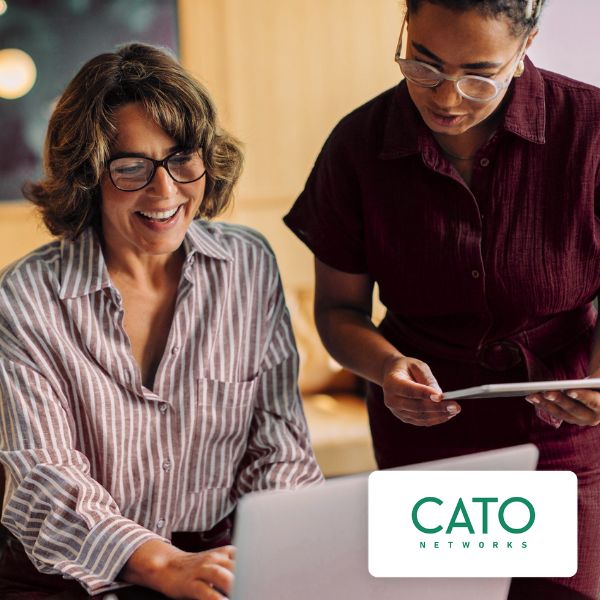 Two women collaborate in an office. One, seated, smiles at a laptop while the other stands, holding a tablet. The mood is friendly and cooperative.