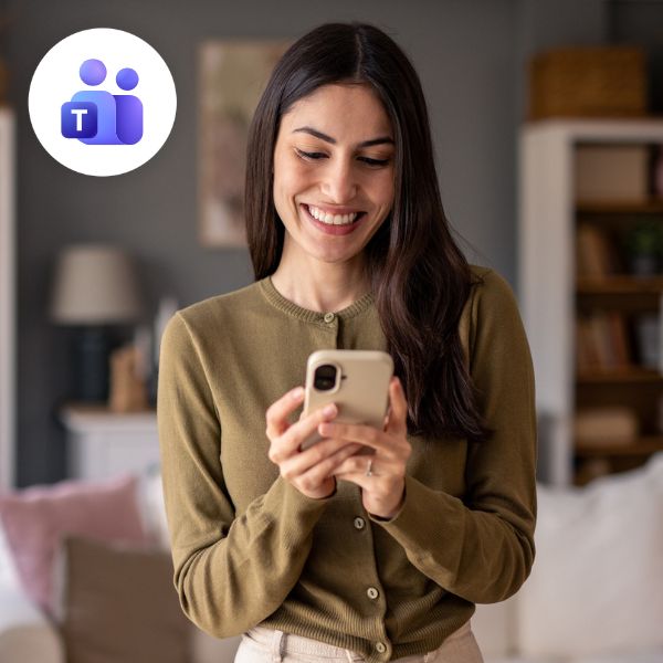 Smiling woman in a cozy room uses a smartphone, with the Microsoft Teams logo in the corner. The scene conveys warmth and connectivity.