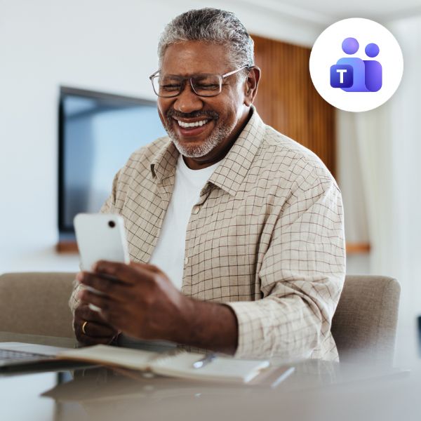 Smiling man with gray hair and glasses, seated, looking at a smartphone. Microsoft Teams logo in the corner, suggesting a virtual meeting. Warm, inviting atmosphere.