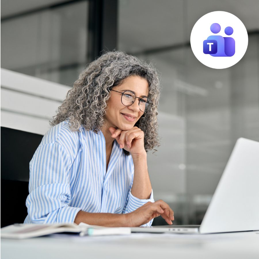 A woman with curly gray hair and glasses smiles while using a laptop. She wears a striped shirt, conveying a professional, relaxed atmosphere. The Microsoft Teams logo is visible in the corner.