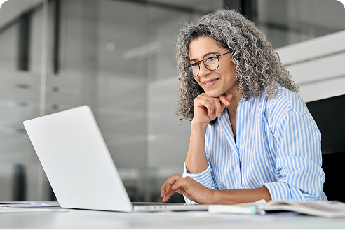 A woman with gray curly hair and glasses, wearing a blue striped shirt, smiles while working on a laptop at a desk in a modern office setting.