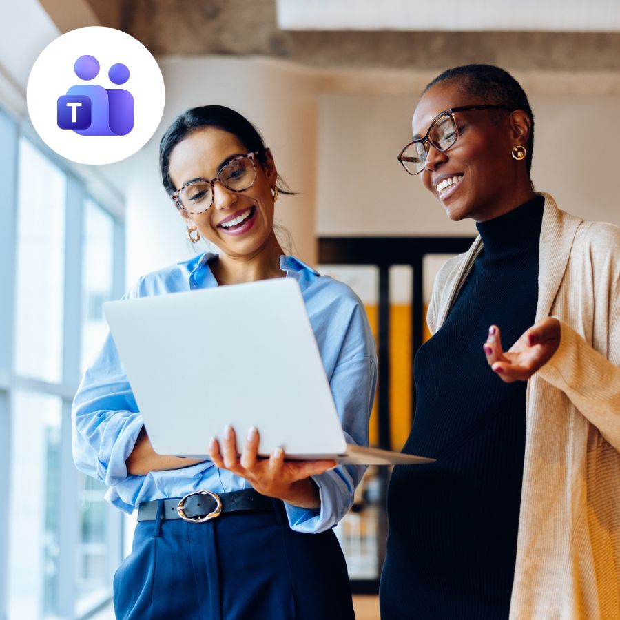 Two women smiling and looking at a laptop in an office. The woman on the left in a blue shirt holds the laptop. A Microsoft Teams logo is in the upper left corner.