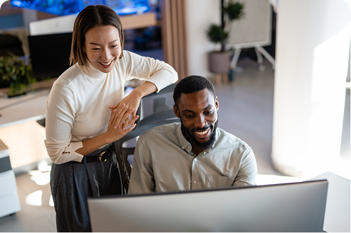 Two people smiling and looking at a computer screen in a modern office. The atmosphere is collaborative and positive, with plants and a flipchart in the background.