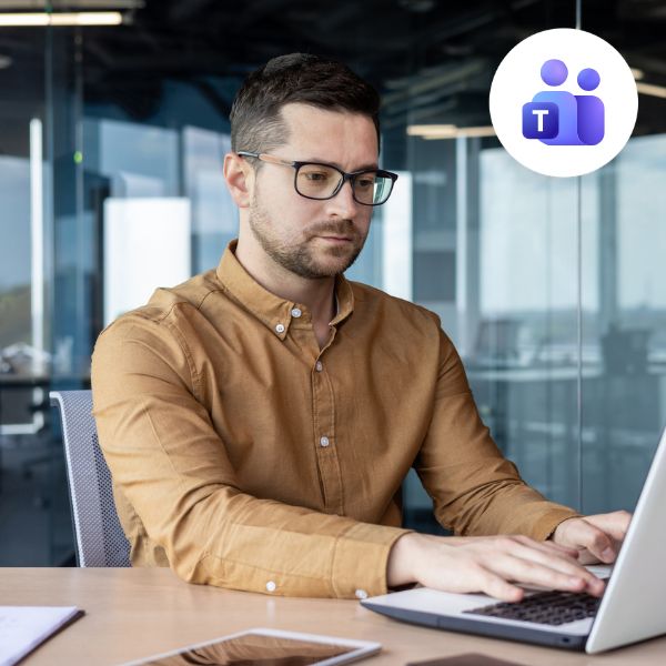 A focused man in a brown shirt and glasses works on a laptop in a modern office with glass walls. Microsoft Teams icon is visible, indicating teamwork.