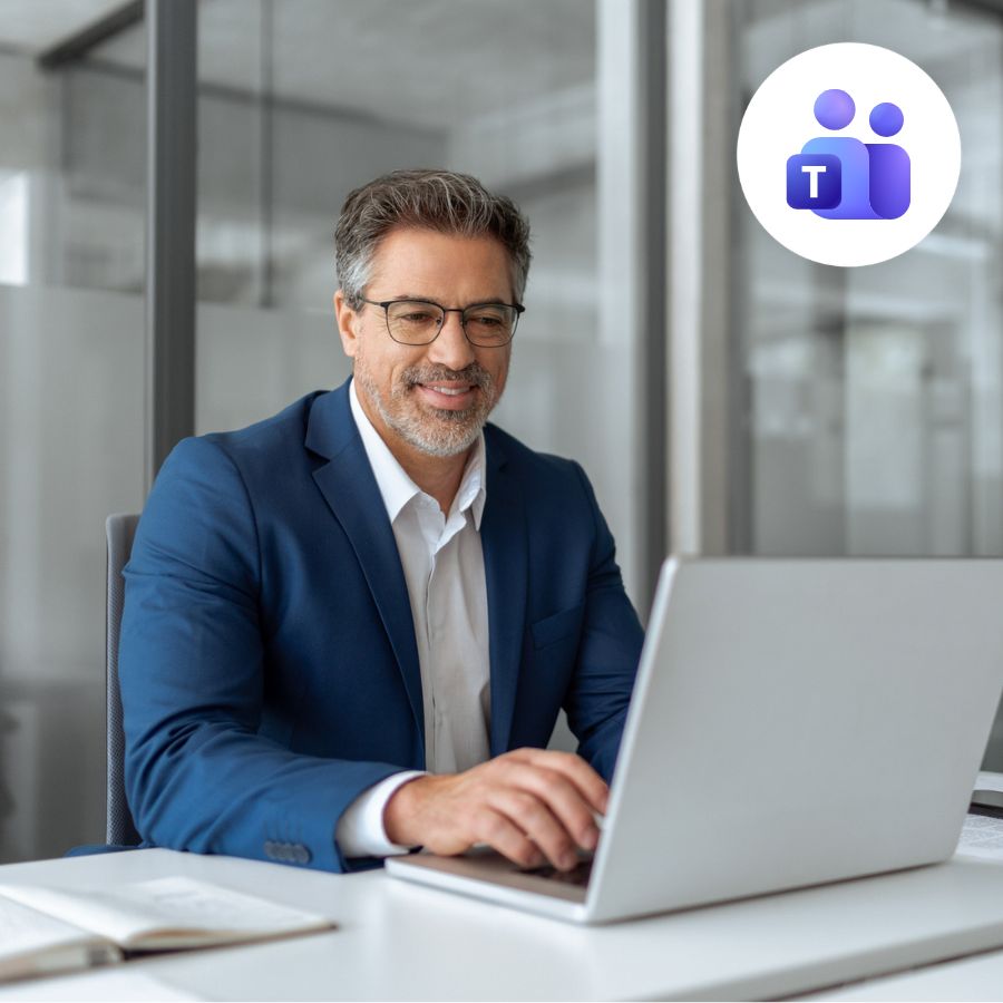 A man in a blue suit and glasses is smiling while typing on a laptop in a modern office. Microsoft Teams logo is visible in the top right corner.
