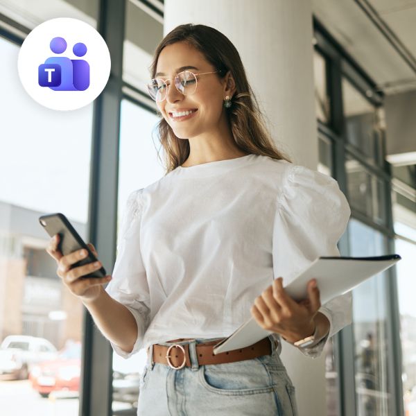 Young woman in a white blouse and jeans, smiling while looking at her phone. She holds a tablet. Microsoft Teams logo in the top left, bright and modern setting.
