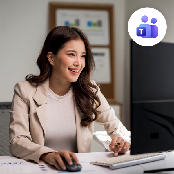 A woman in a beige blazer smiles while using a computer, conveying a positive and engaged mood. Office background with a Microsoft Teams icon visible.