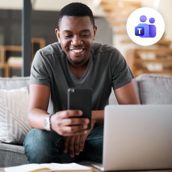 Smiling man in a gray t-shirt using a smartphone, seated on a couch with an open laptop. Microsoft Teams logo in the corner. Relaxed and connected.