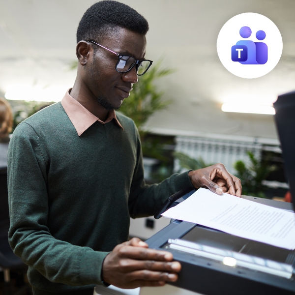 Man in glasses using a photocopier in an office. He is wearing a green sweater with a peach collar. The Microsoft Teams logo is at the top right corner.
