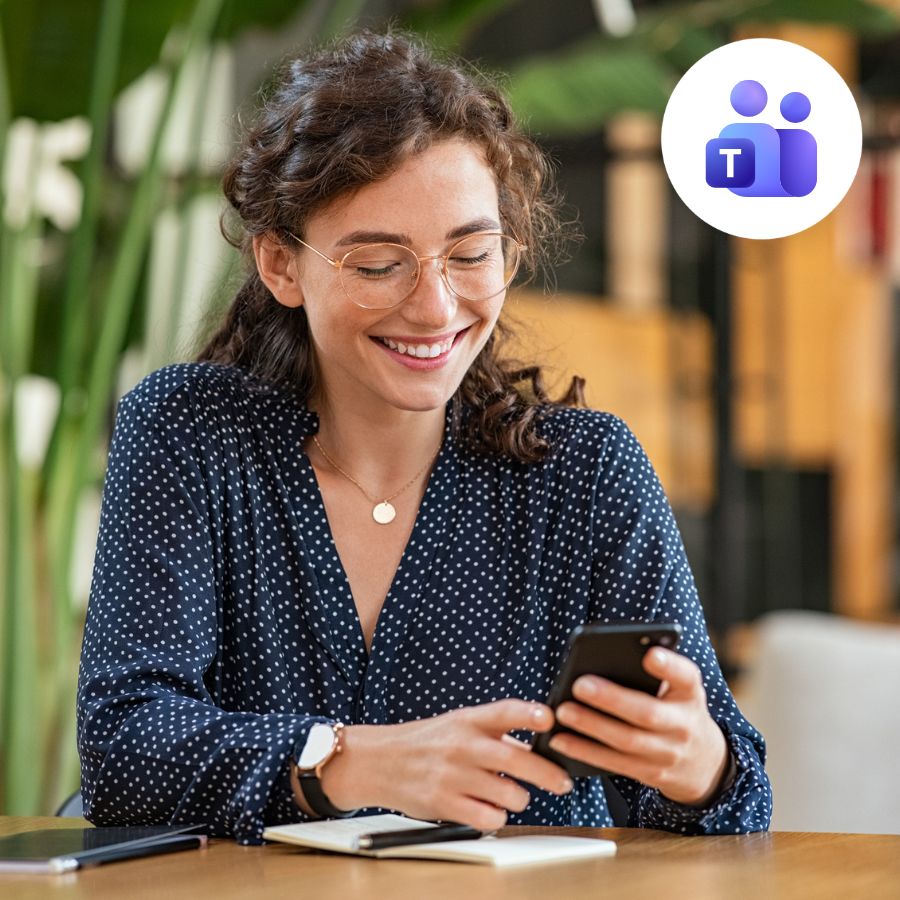 Smiling woman in glasses using a smartphone at a desk with a notebook. Microsoft Teams icon in the corner, suggesting communication. Bright, cheerful setting.
