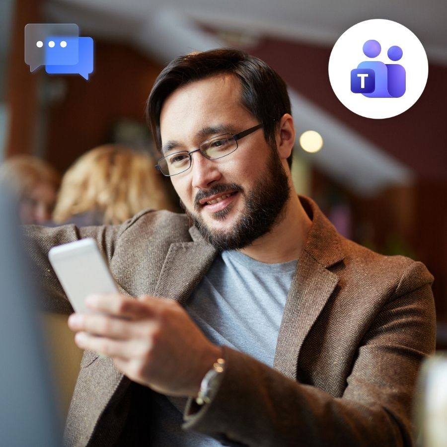Bearded man in a brown blazer smiling at a text message in a café, Teams and chat icons.